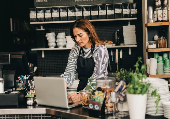 A café owner stands at the counter working on her laptop